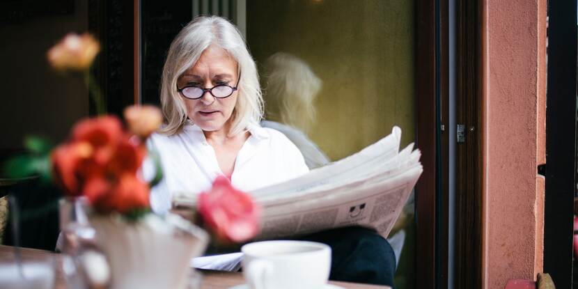 Oudere vrouw met bril leest de krant bij een kopje koffie